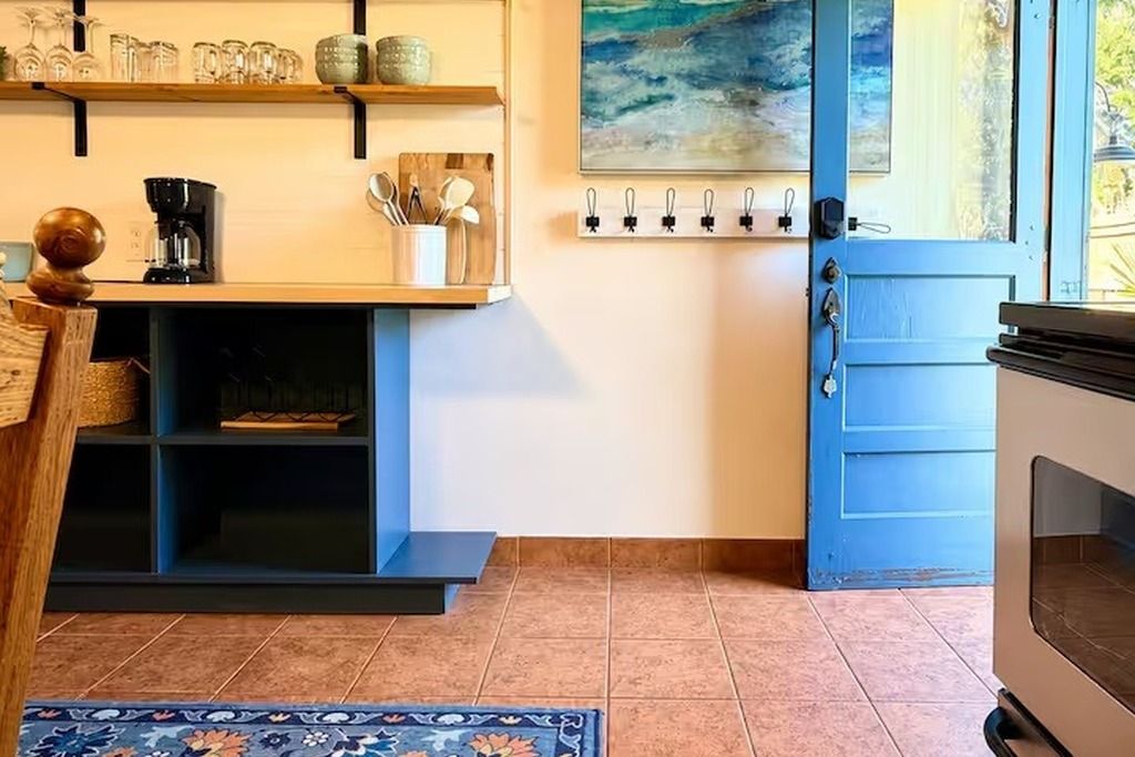A kitchen with a coffee maker, shelves with glassware, a blue door, and a painting on the wall.
