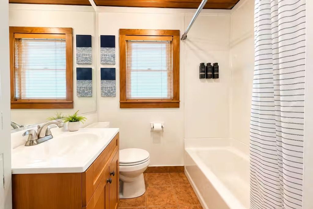 Un baño con un lavabo blanco, tocador de madera, dos ventanas con persianas, inodoro y bañera.