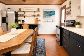 Una cocina con una mesa de comedor de madera, sillas, un refrigerador, una cafetera y una estufa.