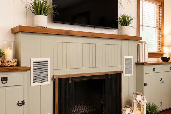 A living room with a fireplace, a television mounted above it, and a window with blinds.