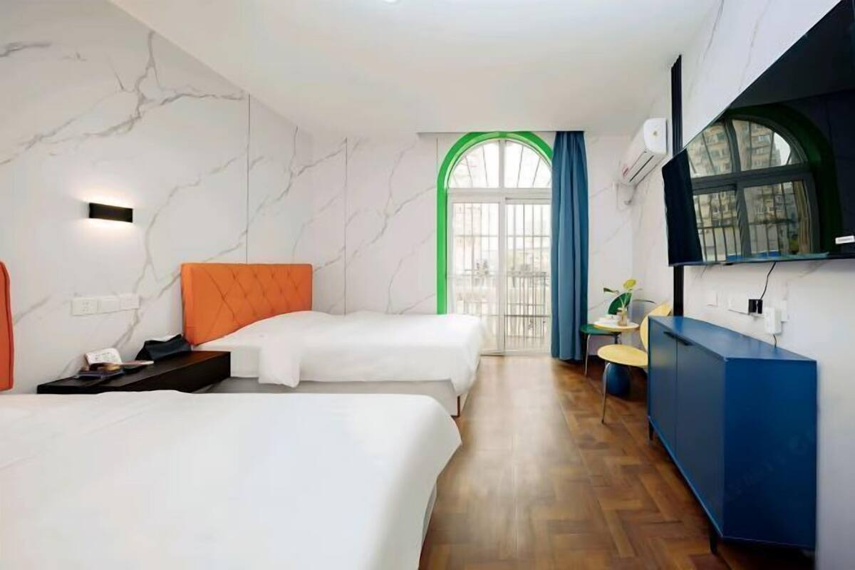 A hotel room with two beds, a desk, a chair, a small table, a window with curtains, and a blue cabinet.