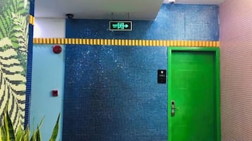A green door with a digital sign above it, a red emergency button, and a patterned wall to the left.