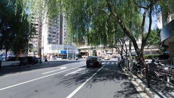 A city street lined with trees, parked cars, and a building with a blue sign.