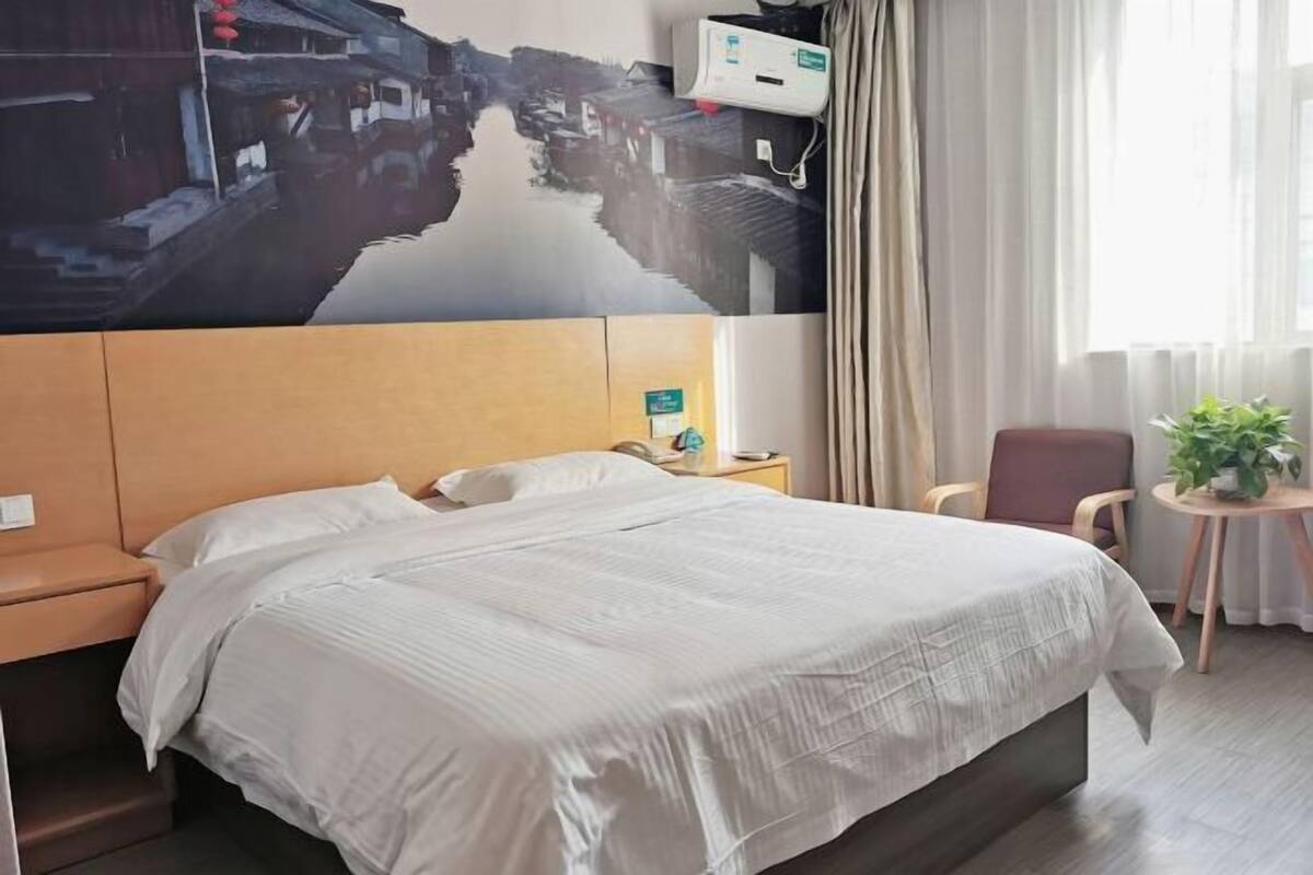 A hotel room with a bed, a nightstand, a chair, a small table with a plant, and a wall-mounted air conditioner.