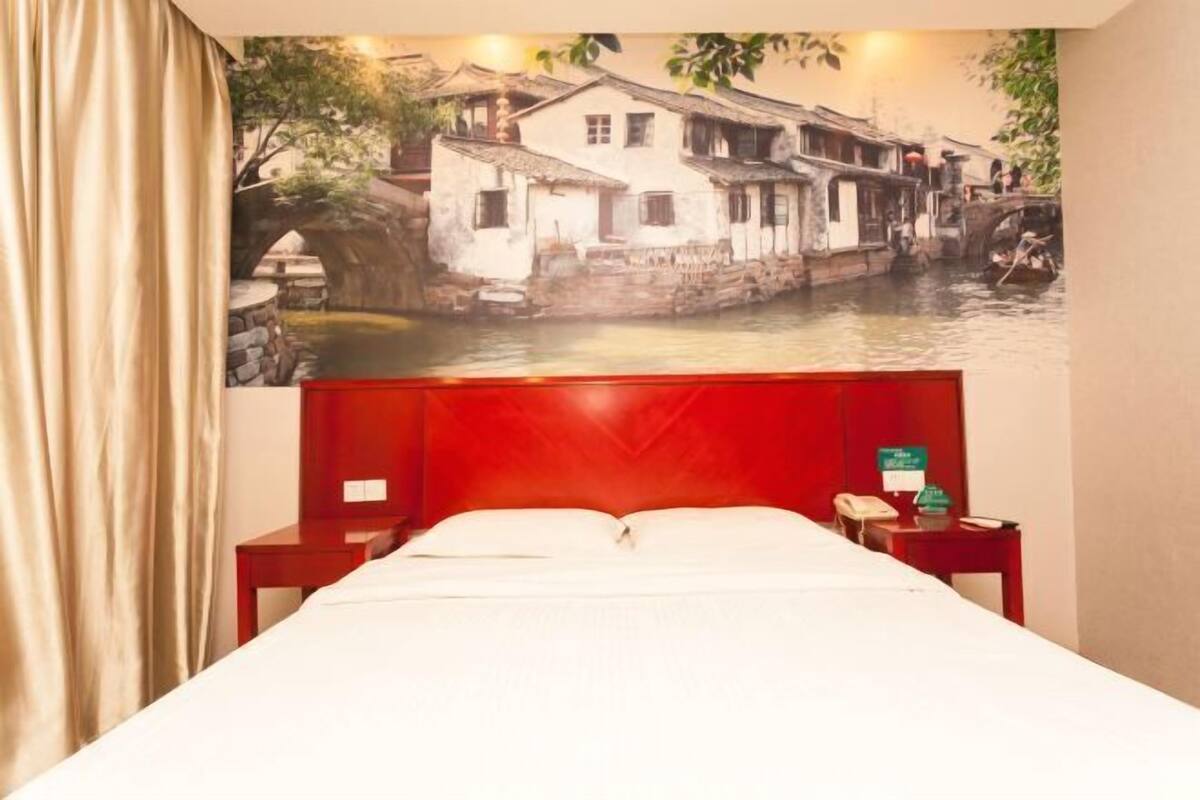 A hotel room with a bed, a red headboard, a nightstand with a telephone, and a wall mural of a traditional building by a waterway.