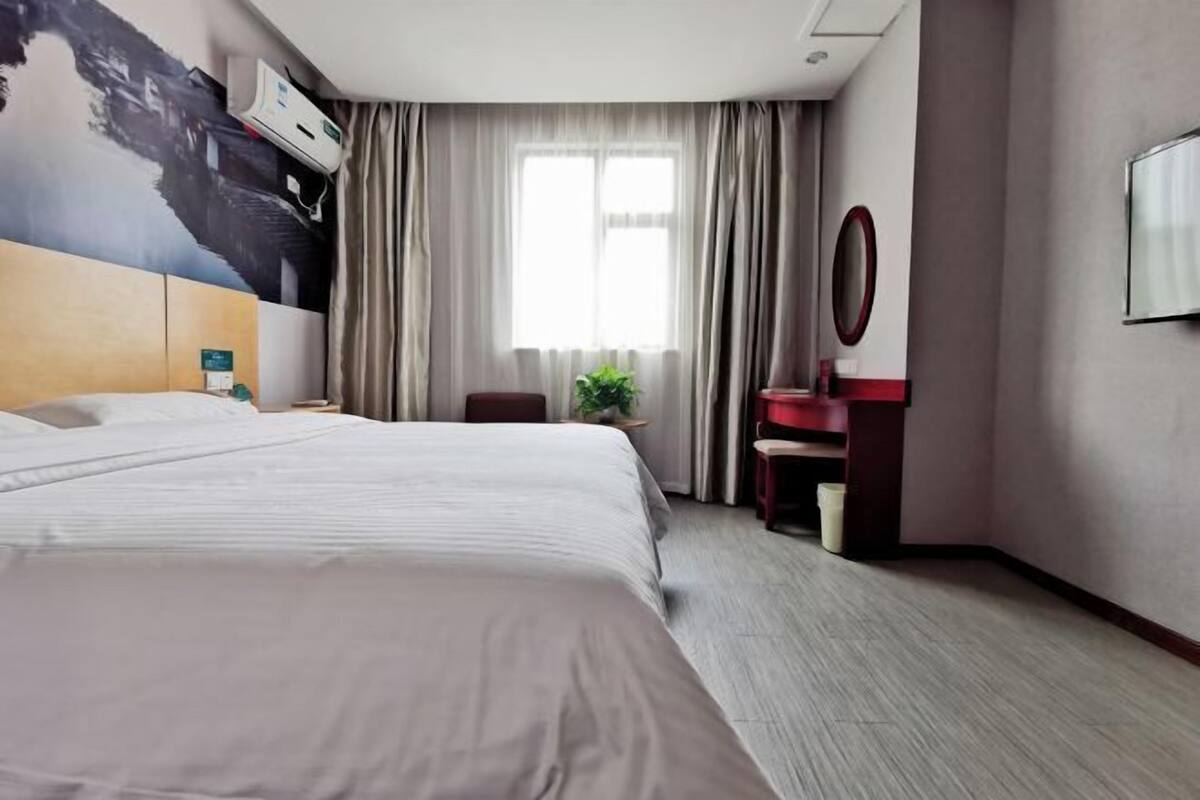 A hotel room with a large bed, a desk with a mirror, a small plant, and a window with curtains.