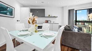 Ein moderner Essbereich mit einem hellblauen Tisch, gedeckt für vier Personen, weißen Stühlen und einer Vase mit Trockenblumen. Im Hintergrund ist eine Küche mit einem großen Fenster zu sehen.
