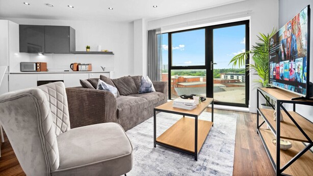 Ein modernes Wohnzimmer mit Sofa, Sessel und Couchtisch. Es gibt eine Küchenzeile mit Mikrowelle und Fernseher. Das Zimmer hat große Fenster mit Blick auf Häuser und eine Pflanze.