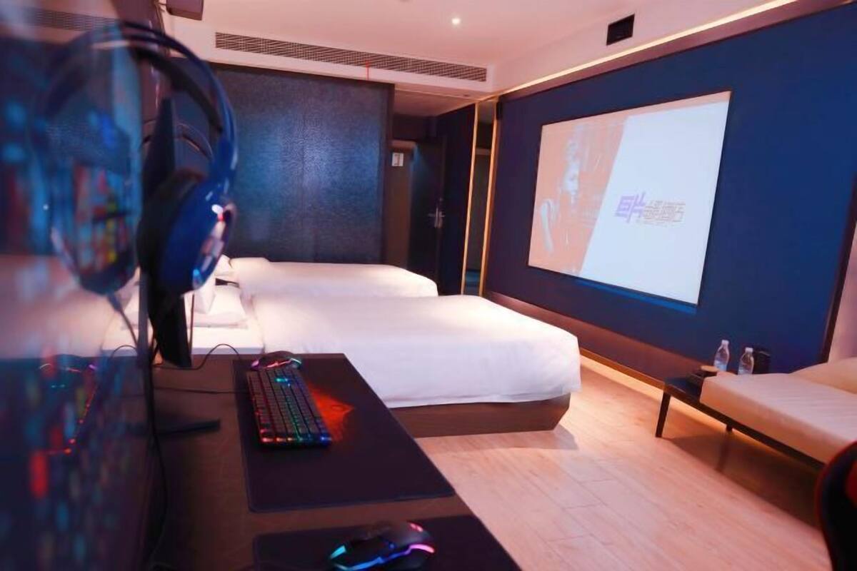 Une chambre d’hôtel équipée d’un lit, d’un bureau avec un ordinateur et des écouteurs, d’un écran de projection et d’une petite table avec des bouteilles d’eau.