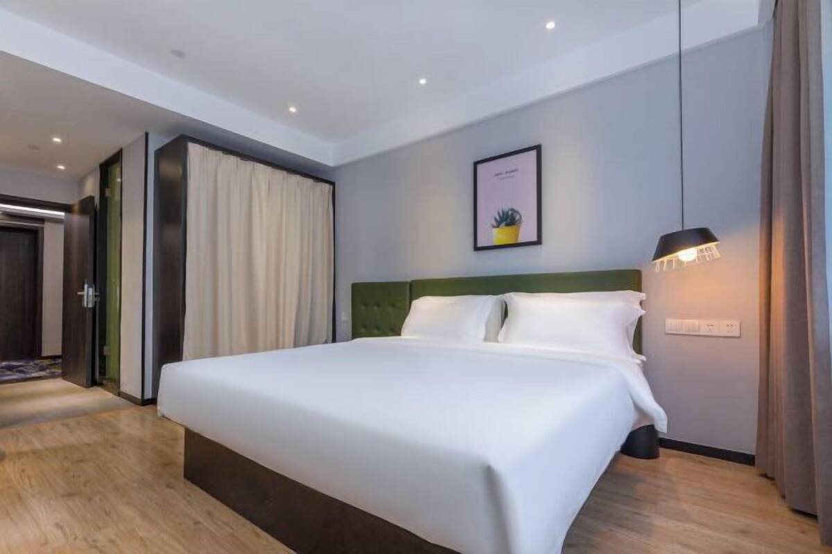 Une chambre d’hôtel moderne avec un grand lit, une tête de lit verte, un tableau encadré au mur et un luminaire suspendu.
