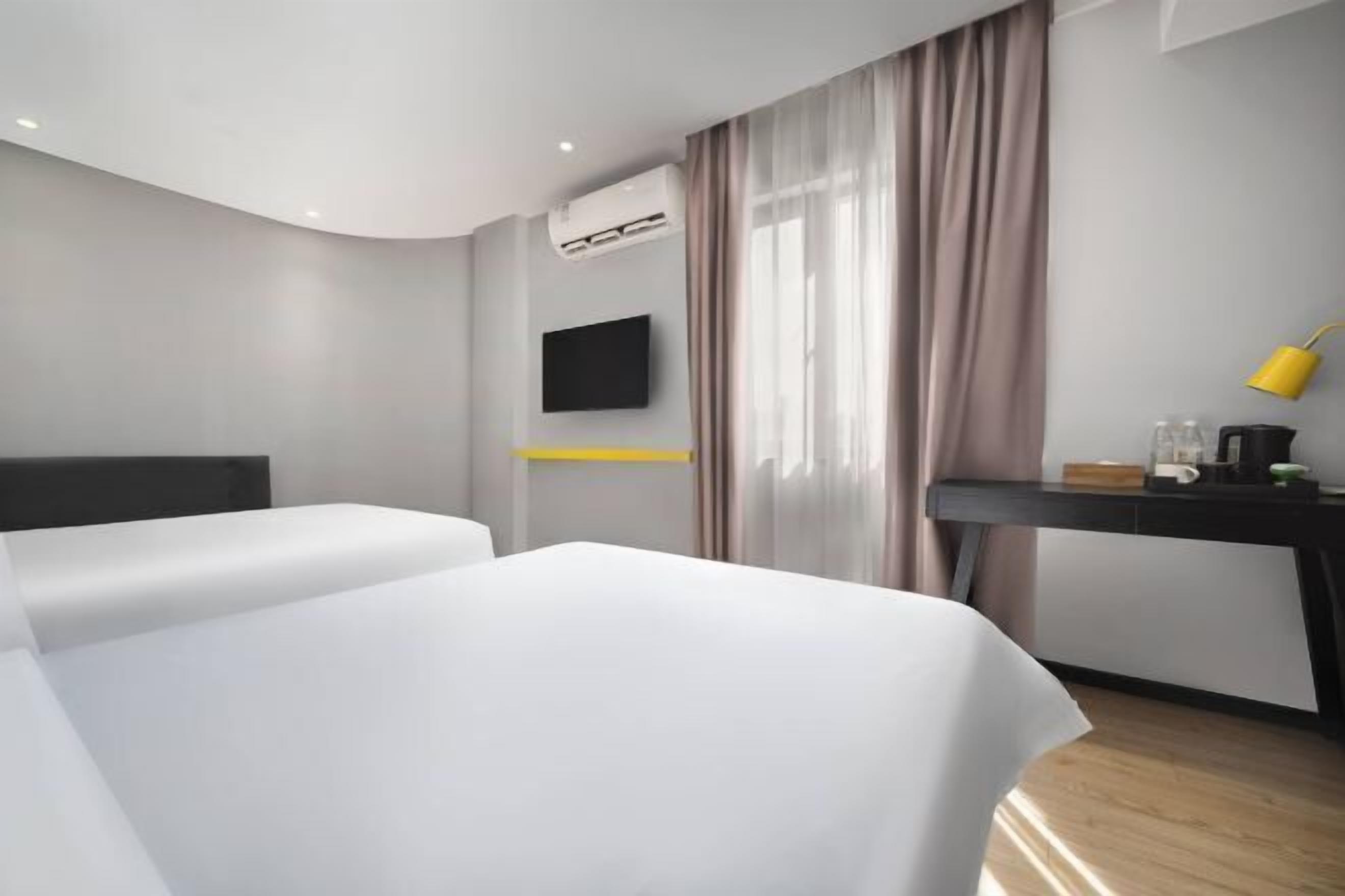 A modern hotel room with a curved ceiling, a flat-screen TV mounted on the wall, a desk with a yellow lamp, and a bed with white bedding.