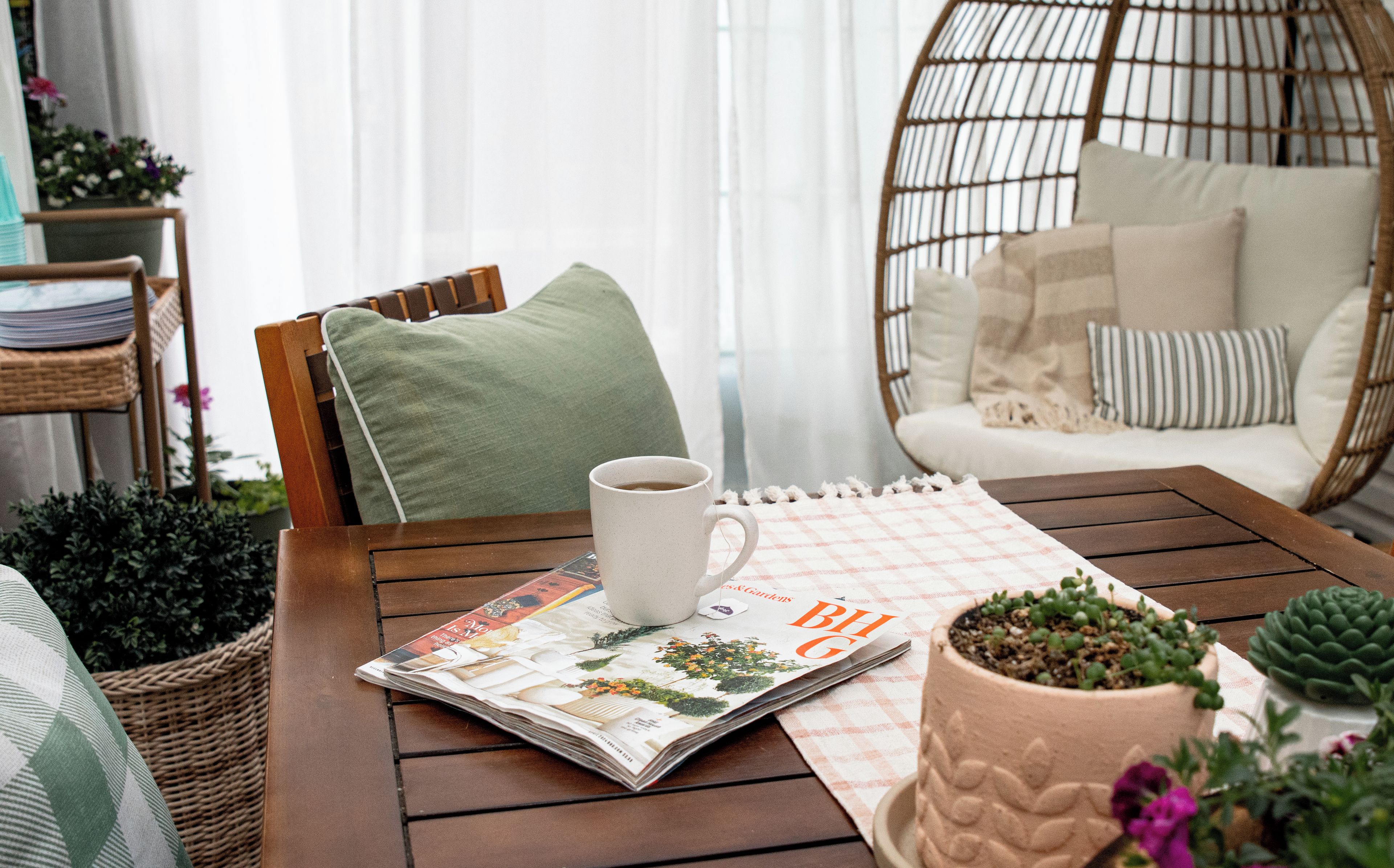 Una silla de mimbre con cojín, una mesa con una revista y una taza, y una planta en maceta.