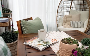 Una silla de mimbre con cojín, una mesa con una revista y una taza, y una planta en maceta.