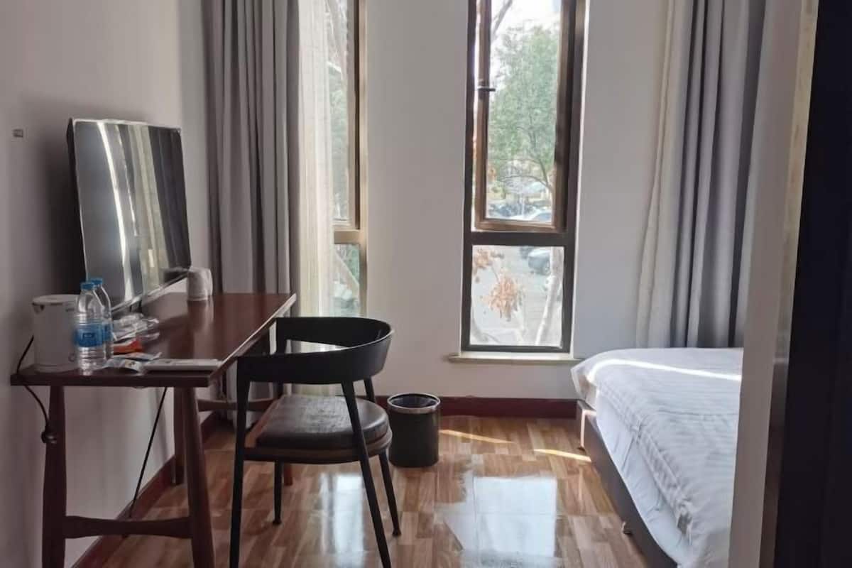 Une chambre d’hôtel comprenant un lit, un bureau avec une chaise, un grand miroir, une fenêtre avec des rideaux et une poubelle.