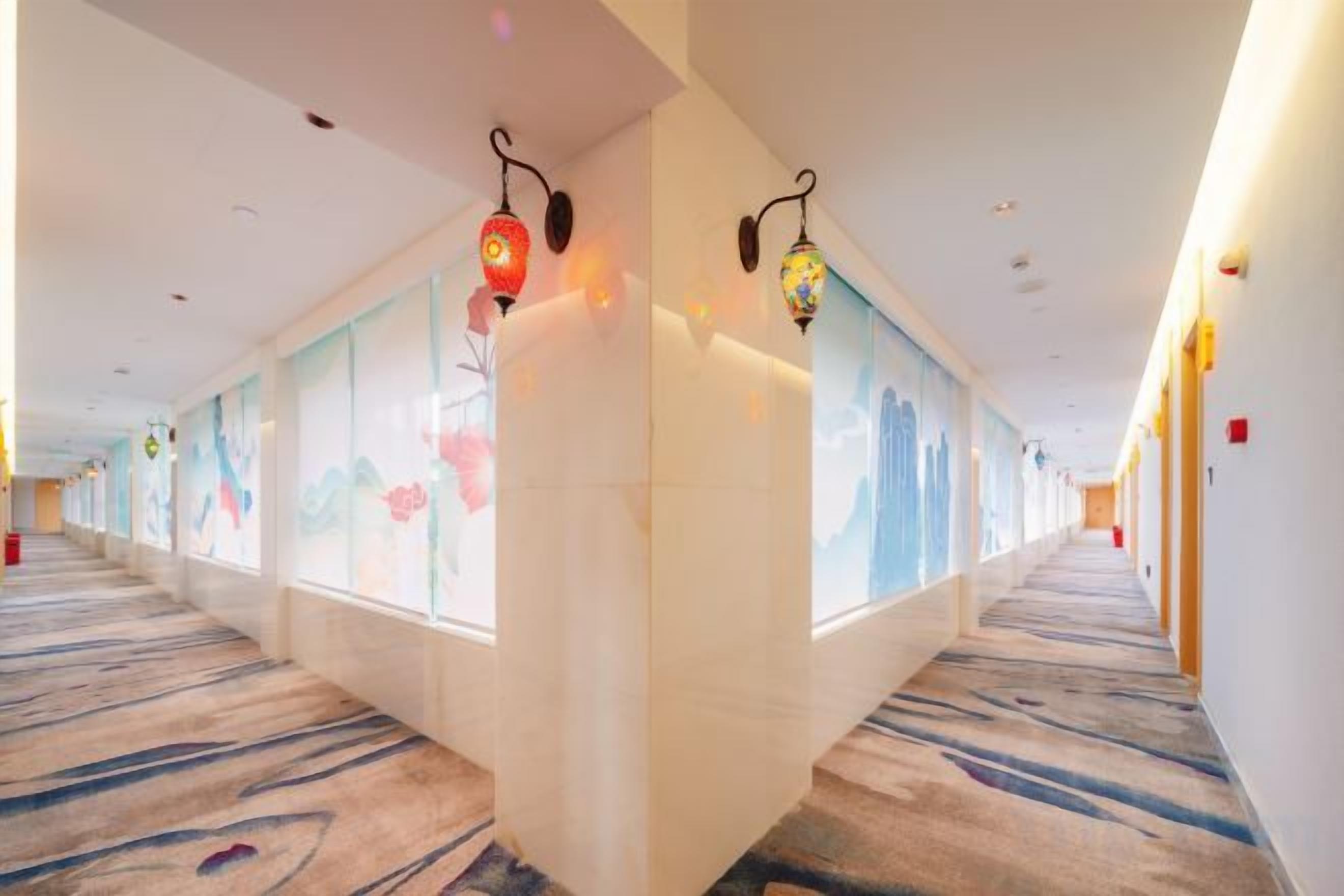 A hotel corridor with a carpeted floor, wall-mounted lamps, and decorative glass panels.