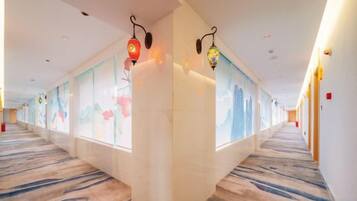 A hotel corridor with a carpeted floor, wall-mounted lamps, and decorative glass panels.