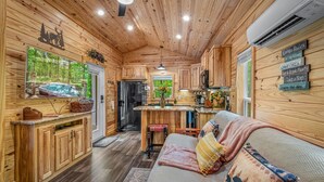 L’intérieur d’une cabane avec des murs en bois, un coin cuisine, un canapé avec des coussins et une grande fenêtre.