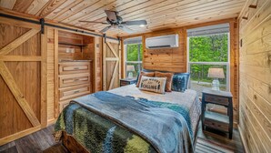 Une chambre dans une cabane en bois, avec un lit, un ventilateur de plafond, une fenêtre donnant sur la forêt et un appareil de climatisation.