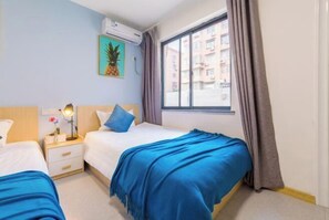 Hotellihuone, jossa on kaksi sänkyä, ikkuna, jossa on verhot, seinälle asennettu ilmastointilaite ja kehystetty taulu.