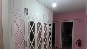 A room with a large white wardrobe with multiple doors and handles, a ceiling light, and a pink wall. - Mehmet Home (Denizli)