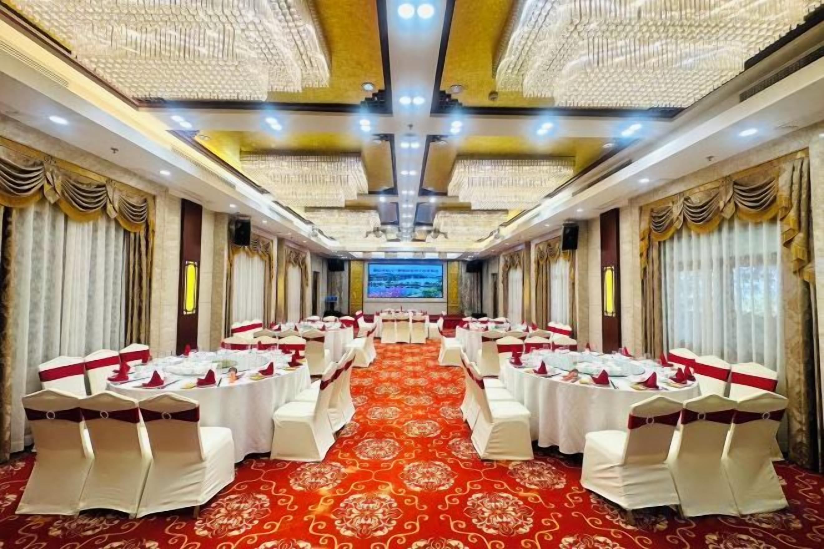Un salón de banquetes con filas de mesas y sillas, una alfombra roja y una pantalla grande al frente.