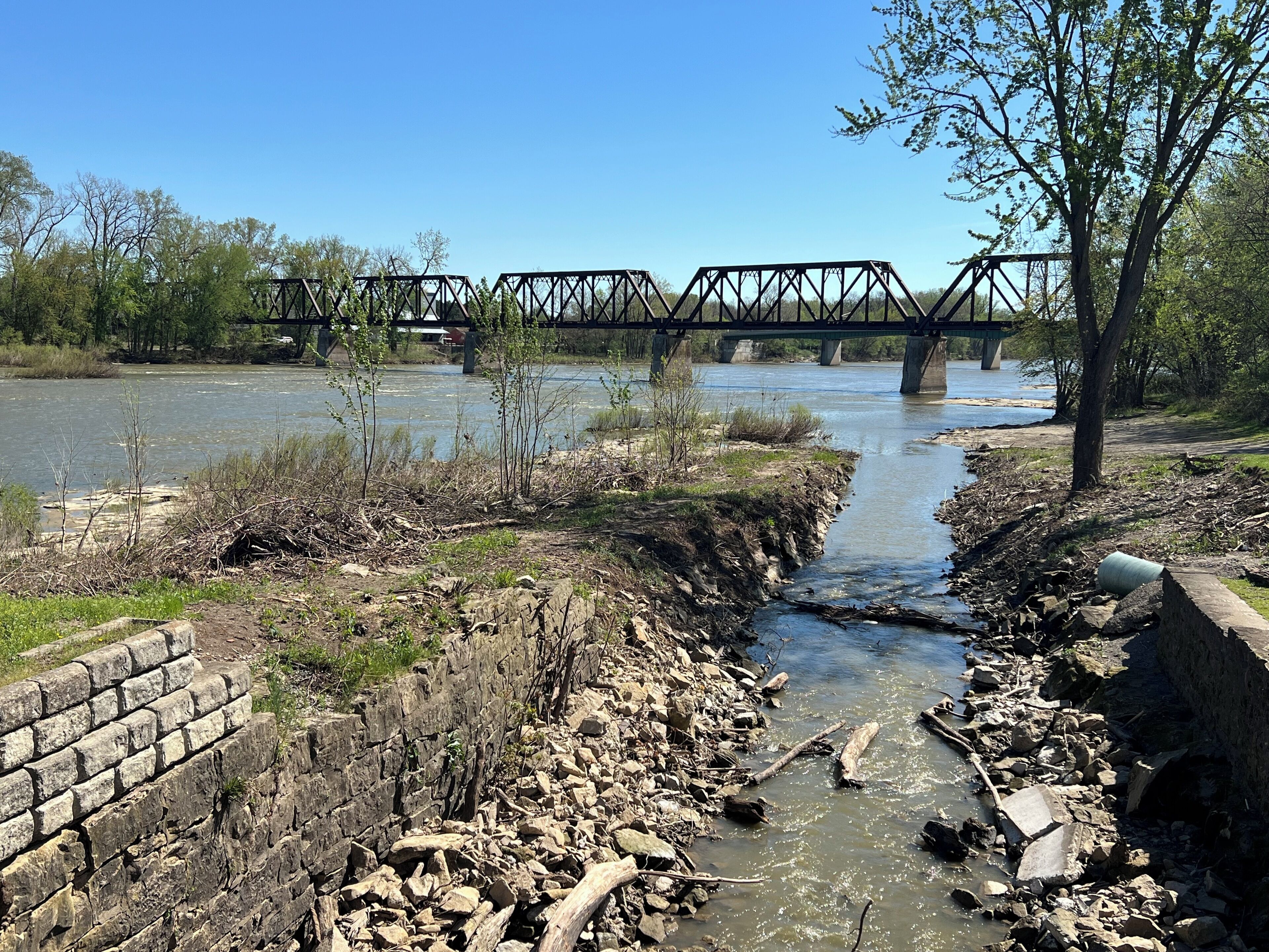 A riverbank with a concrete retaining wall, a metal bridge, and a tree.