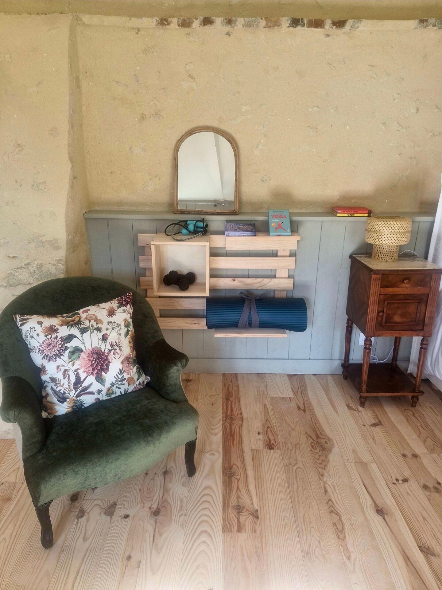 A room with a green armchair, a wooden shelf with books and a mirror, and a small wooden table.
