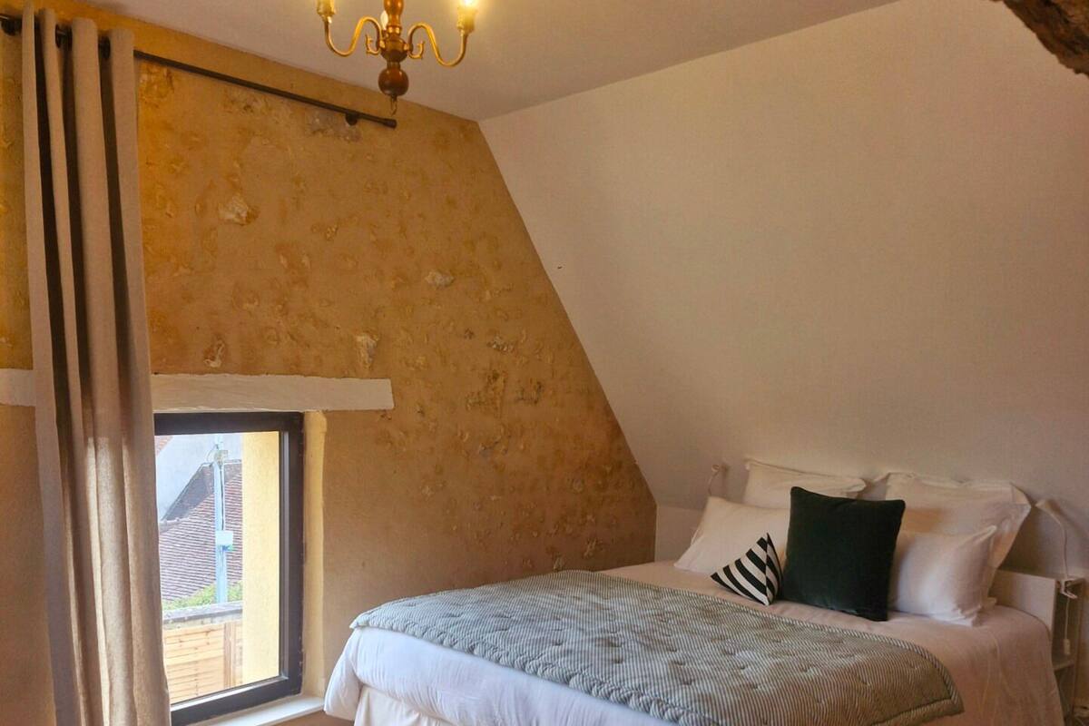 A bedroom with a sloped ceiling, a bed with white bedding, a chandelier, and a window with curtains.