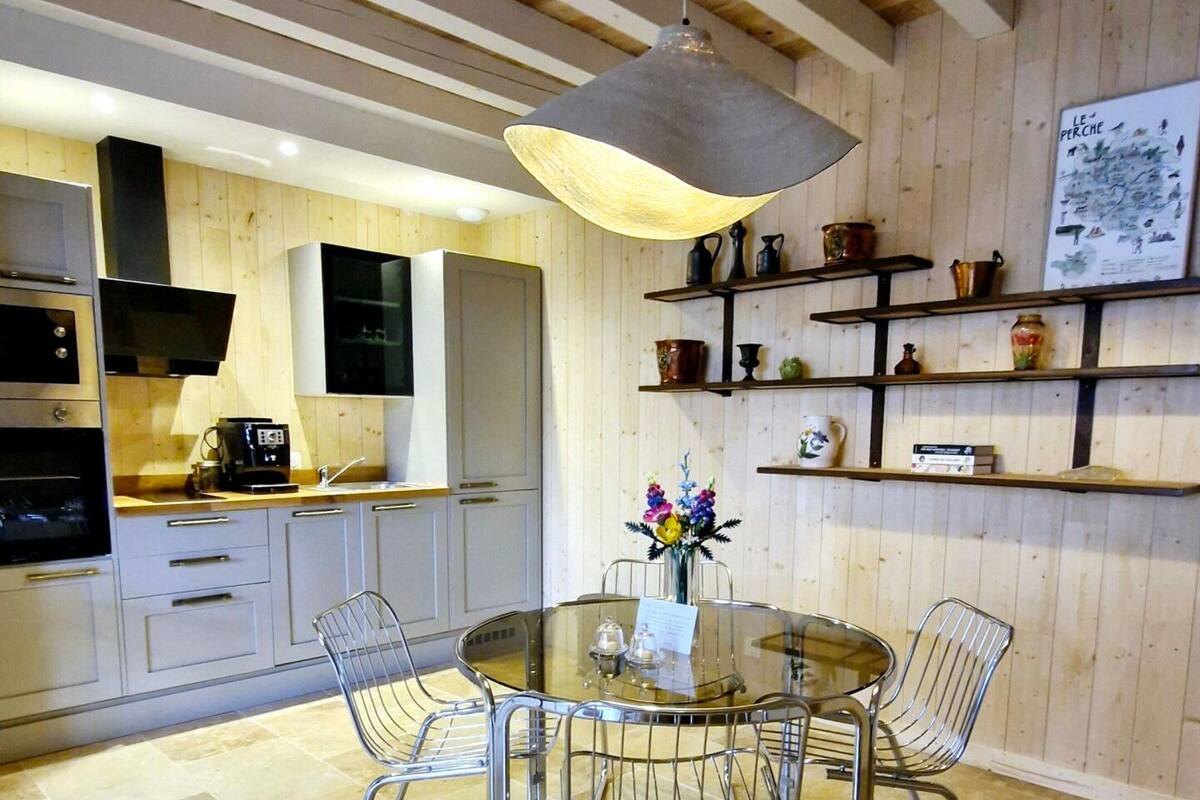 A modern kitchen with a round glass table and metal chairs, wooden beams on the ceiling, and shelves with decorative items.