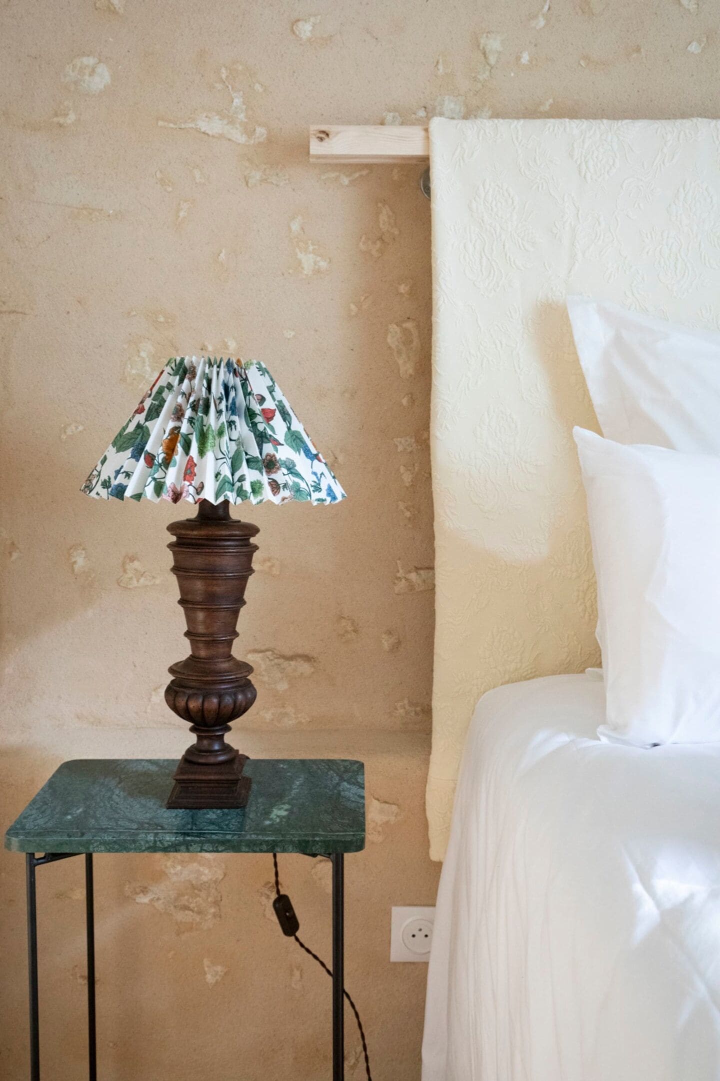 A room with a bed, a small table with a lamp, and a wall with a floral pattern.