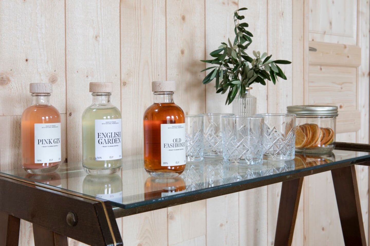 A glass table with three bottles of gin, a vase with green foliage, and a jar of dried orange slices.