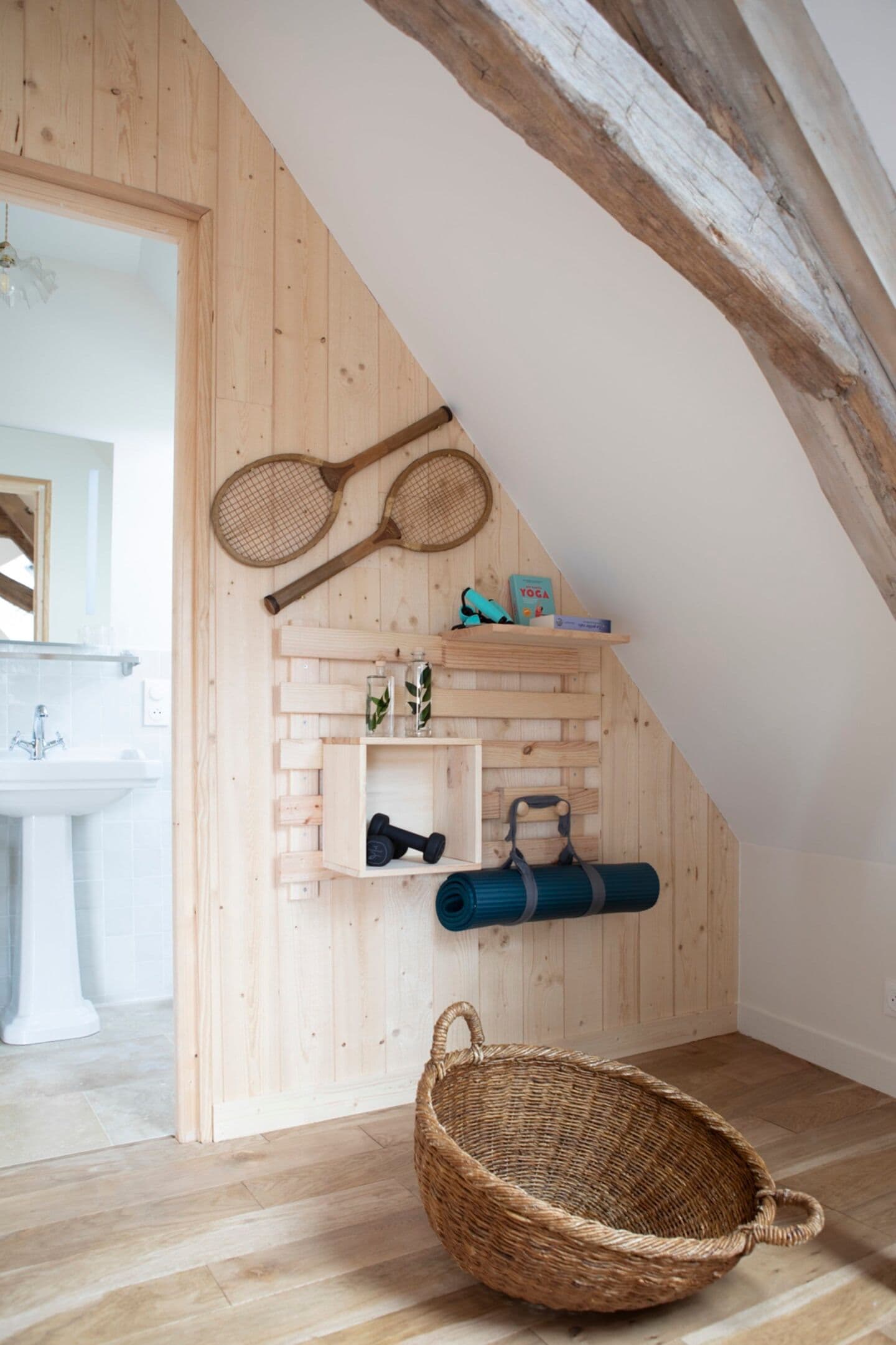 A wooden interior with a sloped ceiling, a wicker basket, shelves with yoga mats, and tennis rackets.