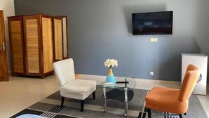 Una sala de estar con pared gris, un televisor de pantalla plana, una mesa de centro de vidrio, dos sillas, un jarrón pequeño con flores y un armario de madera.