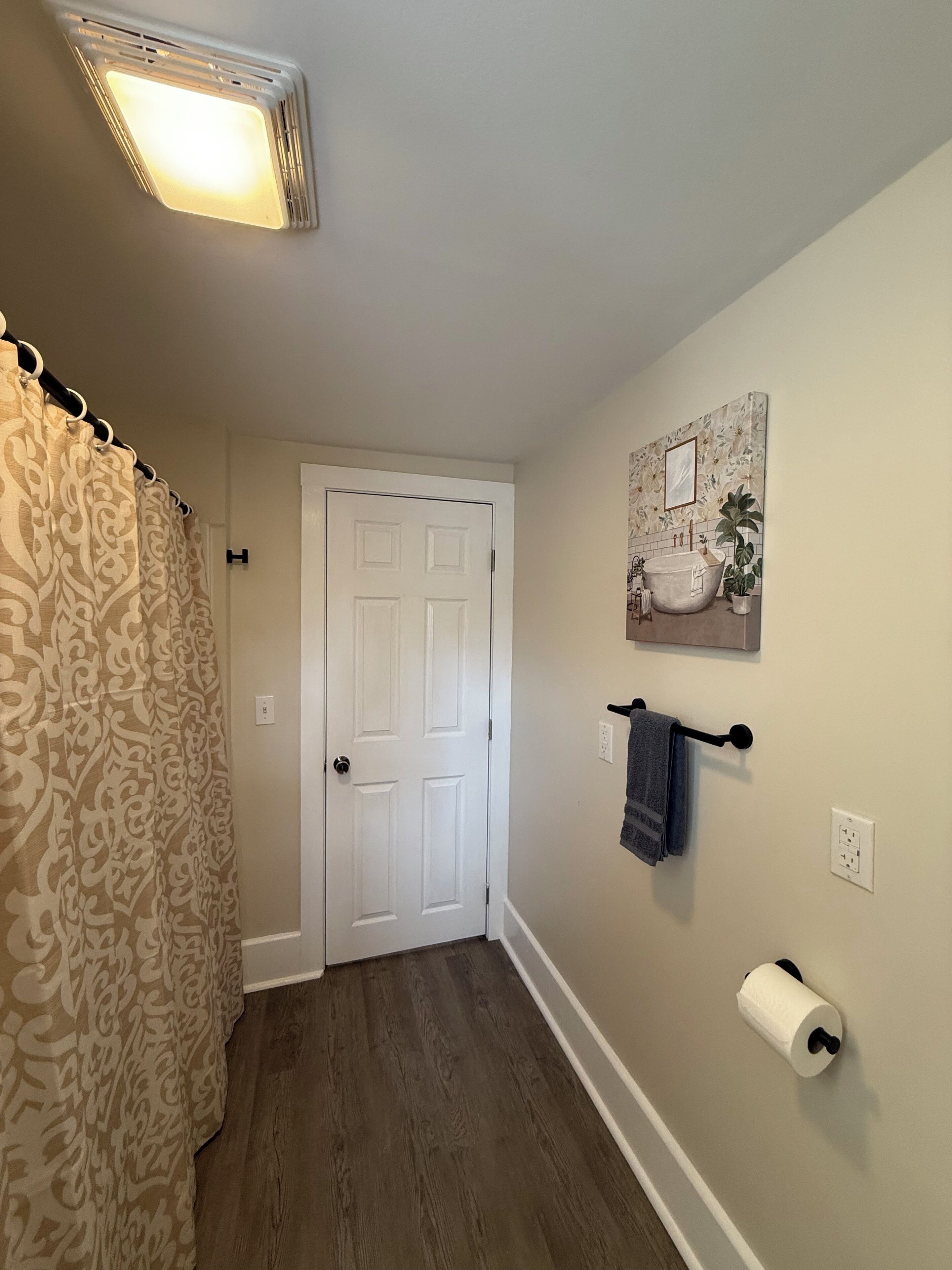 Ein Flur mit einer weißen Tür, gemusterten Vorhängen, einem an der Wand befestigten Handtuchhalter, einem gerahmten Bild und einem Toilettenpapierhalter.