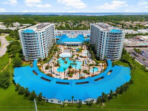 A hotel with a central pool area surrounded by multiple buildings, a large parking lot, and a view of the sea.