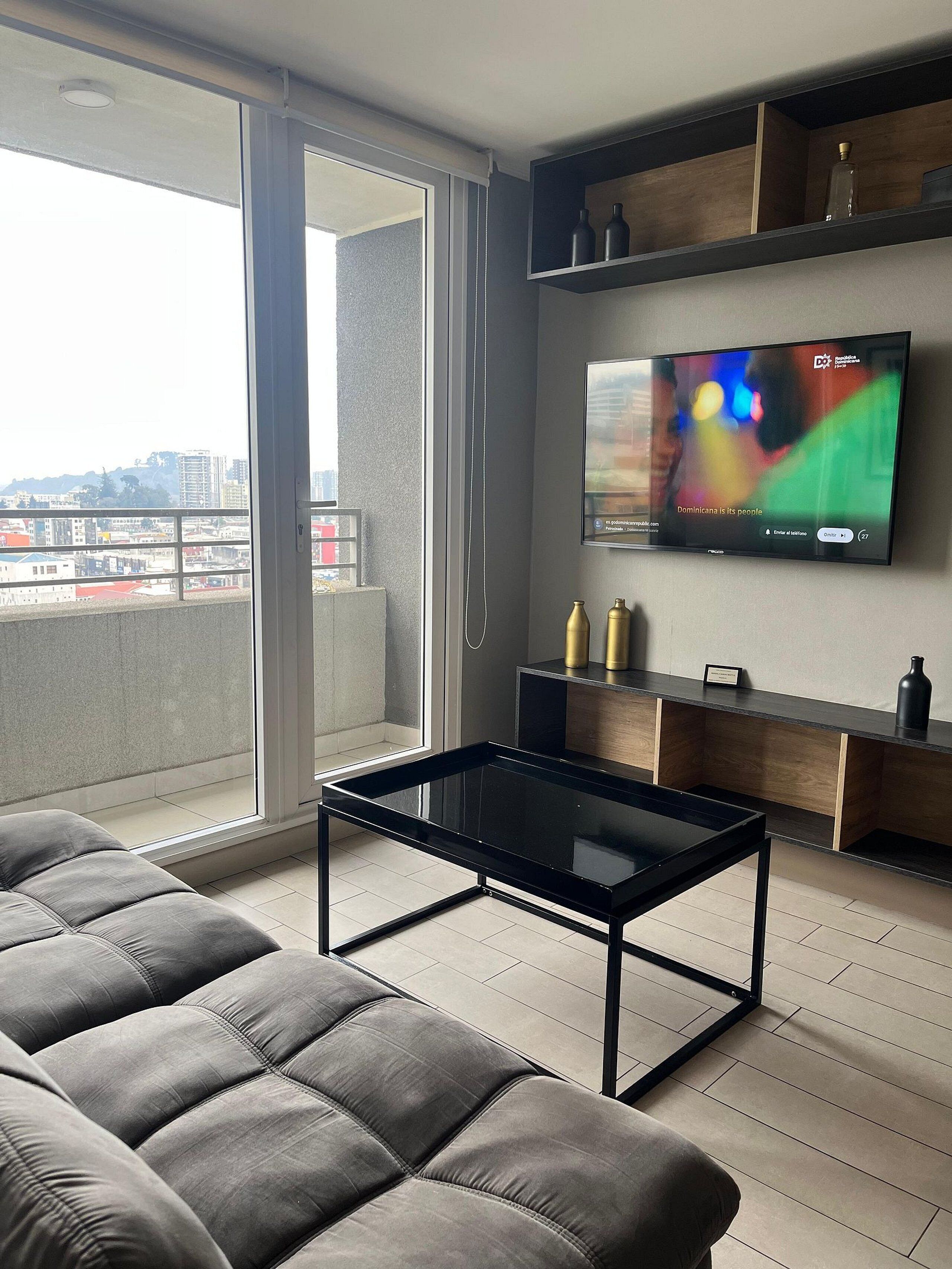 Un salon moderne avec une table basse en verre noir, un canapé gris et un téléviseur à écran plat fixé au mur.