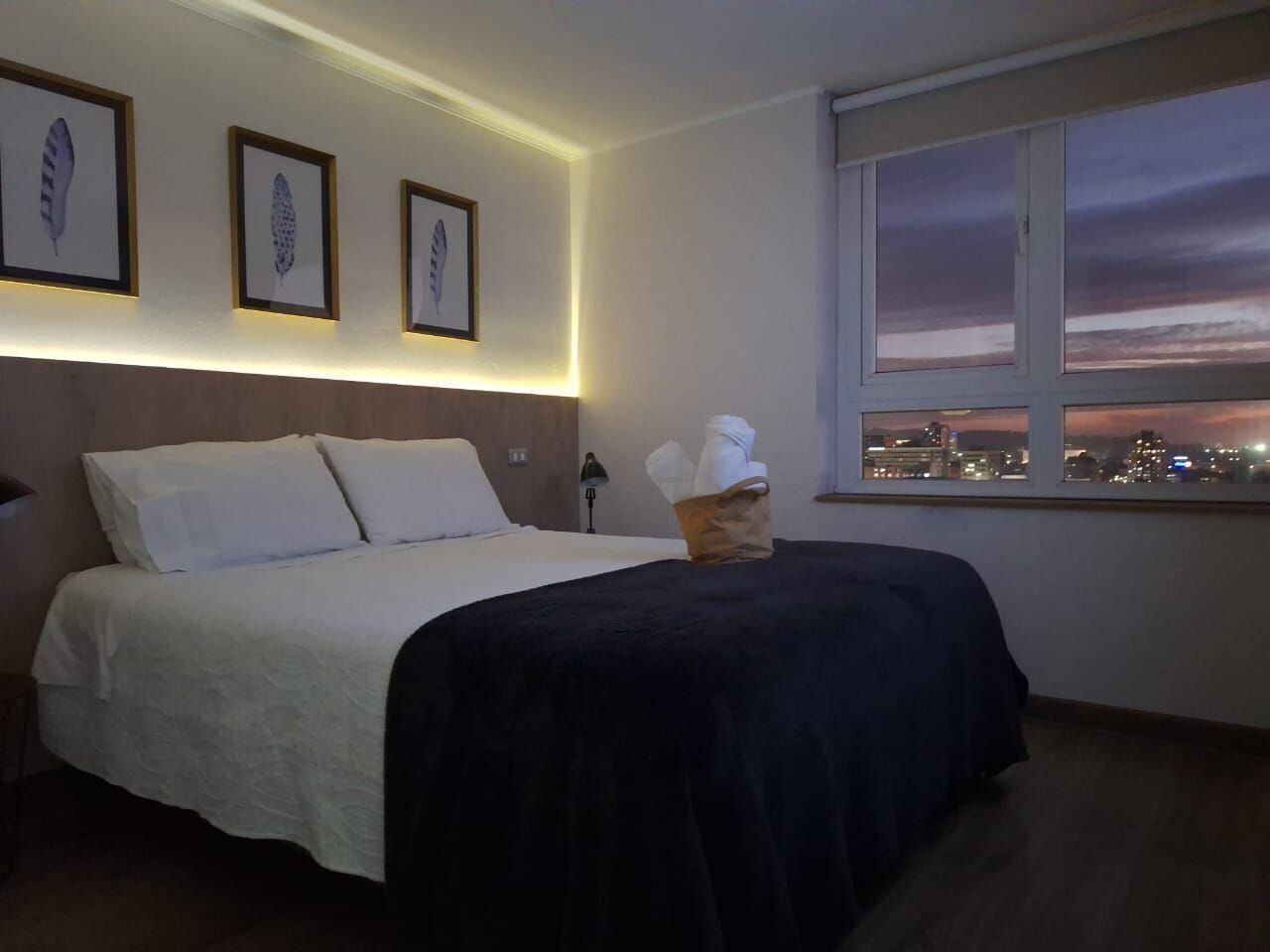 Habitación de hotel con cama, mesita de noche, lámpara, cuadros en la pared y ventana con vista a la ciudad.
