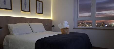 A hotel room with a bed, a nightstand, a lamp, framed artwork on the wall, and a window with a city view.