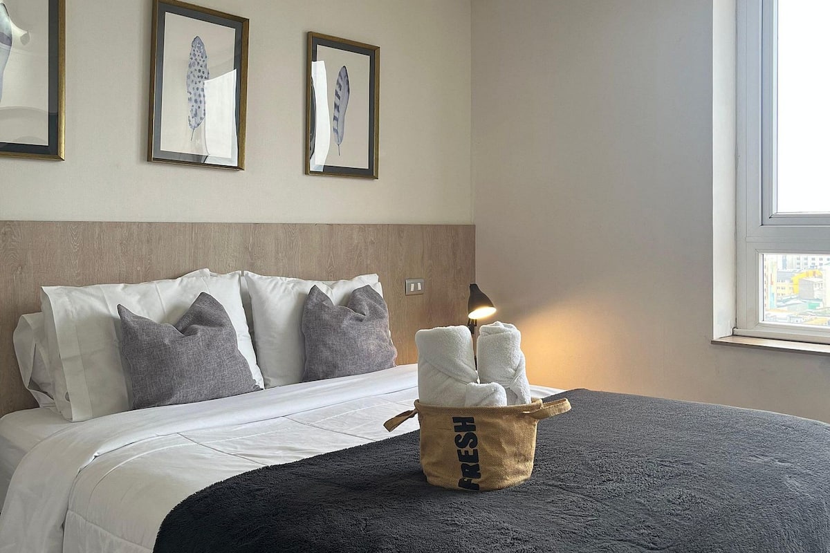 A neatly made bed with a black blanket, a basket labeled "FRESH" with towels, and two framed wall art pieces.