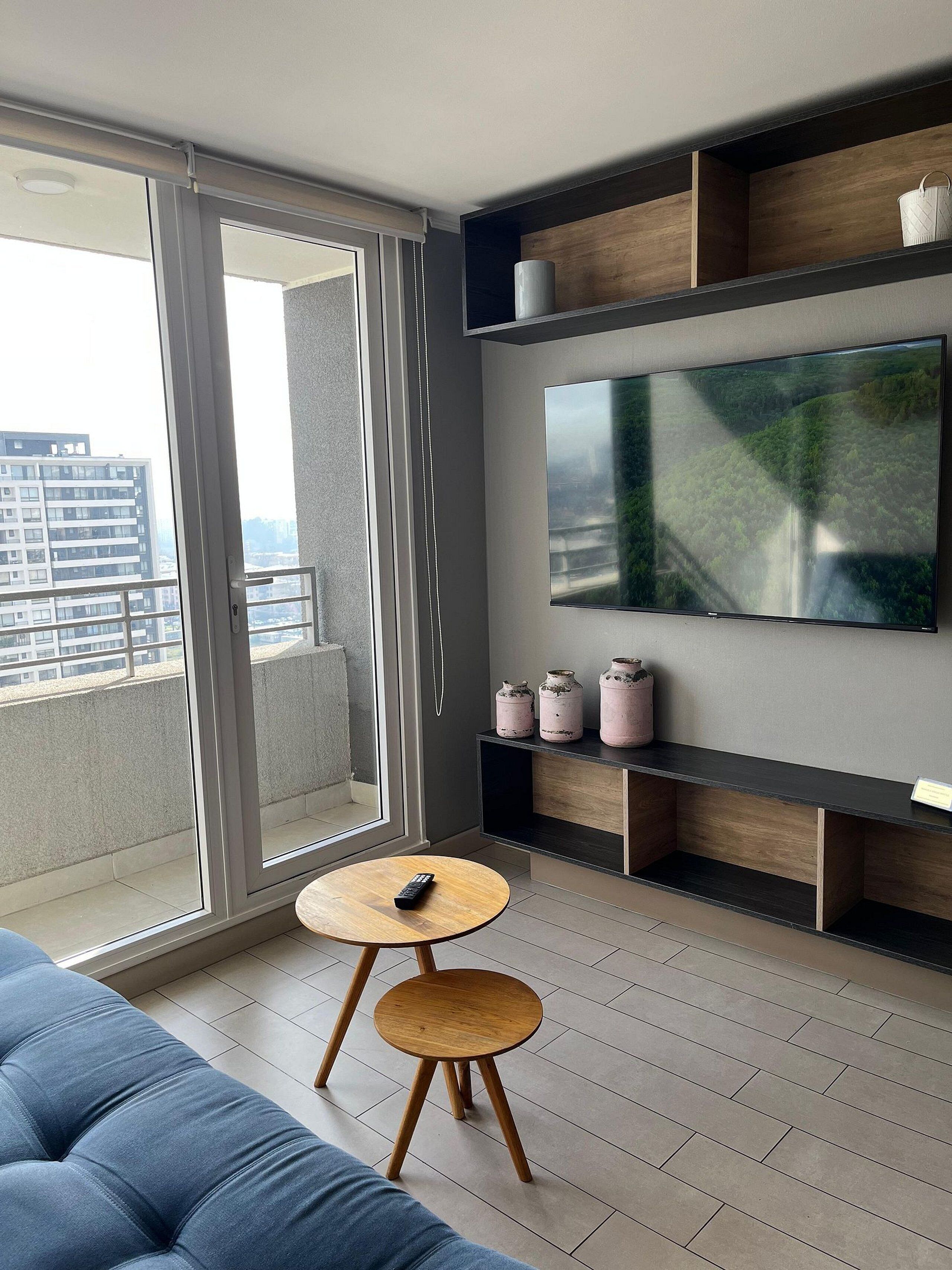 Una sala de estar moderna con un televisor grande, un sofá, una mesita redonda y un ventanal corredero que da a un balcón.