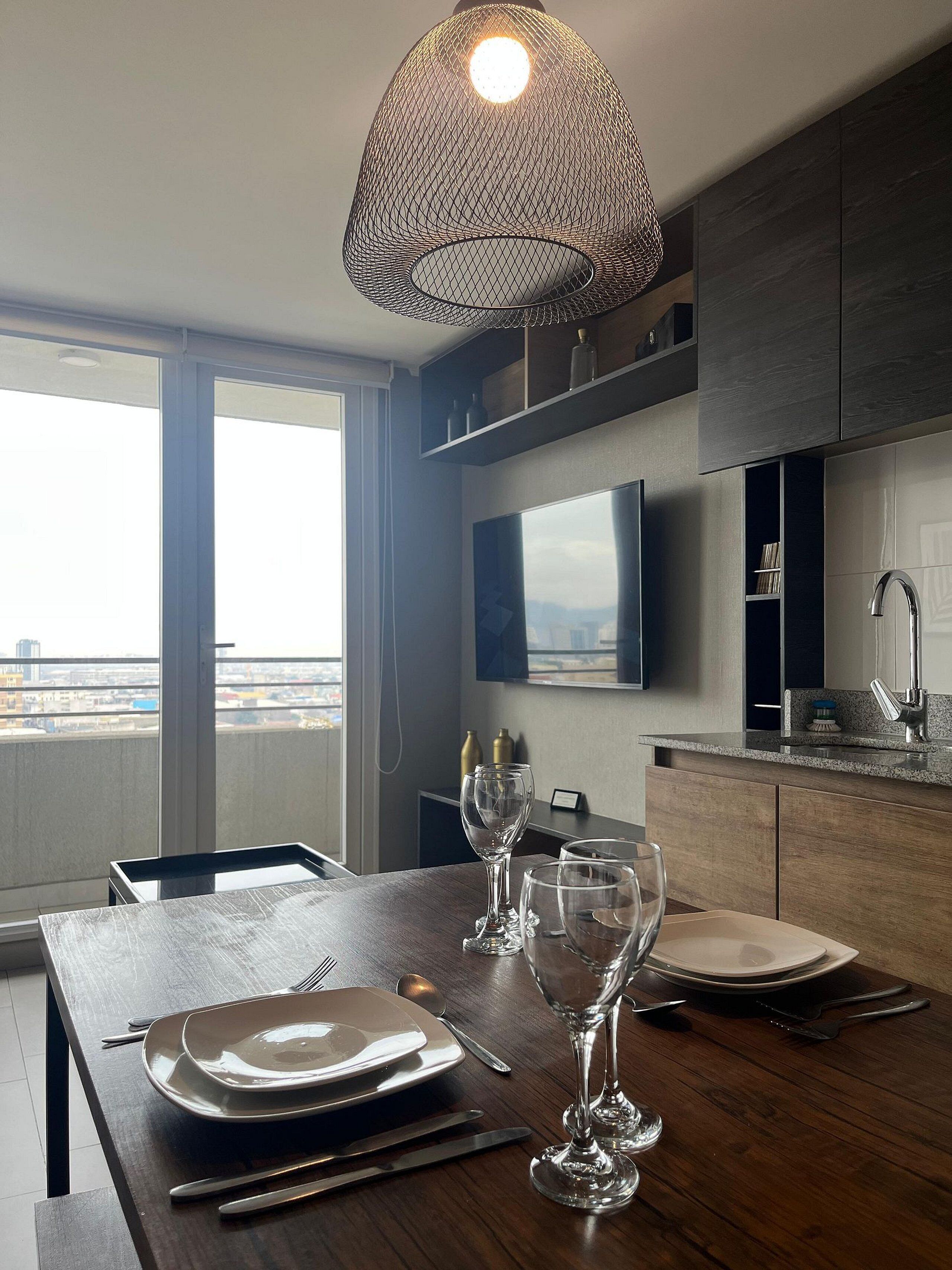 Un comedor moderno con una mesa de madera preparada para cuatro comensales, una lámpara colgante y vistas a la ciudad a través de amplias ventanas.