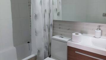 A bathroom with a toilet, a bidet, a shower curtain, a sink with a mirror, and a toilet paper holder.