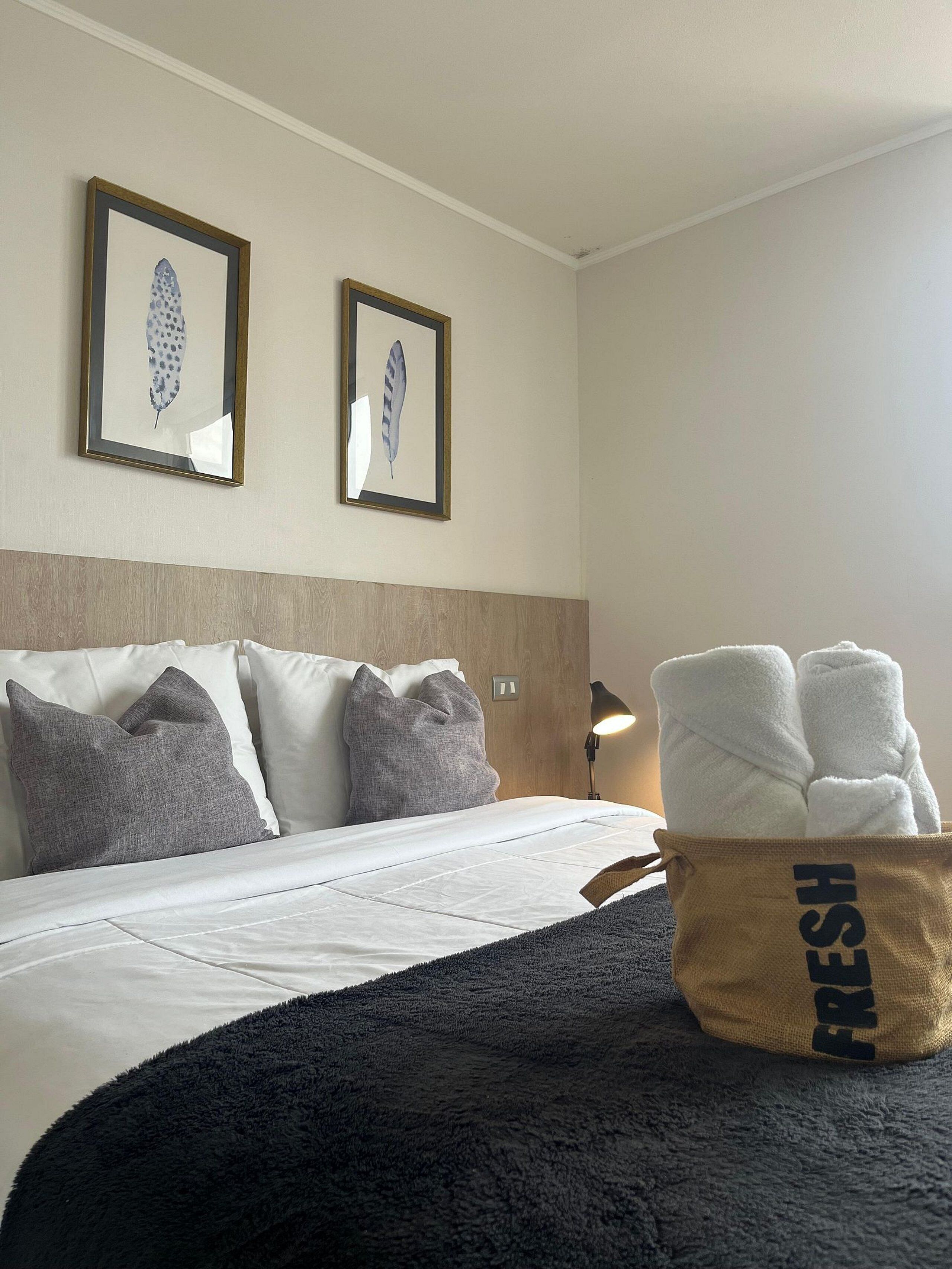 Ett hotellrum med en säng, kuddar, en sänggavel, en lampa och en väska med texten ”FRESH” på, som står på golvet.