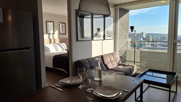 Ein modernes Hotelzimmer mit Balkon, einem für zwei Personen gedeckten Esstisch und einem Bett mit Blick auf die Stadt.