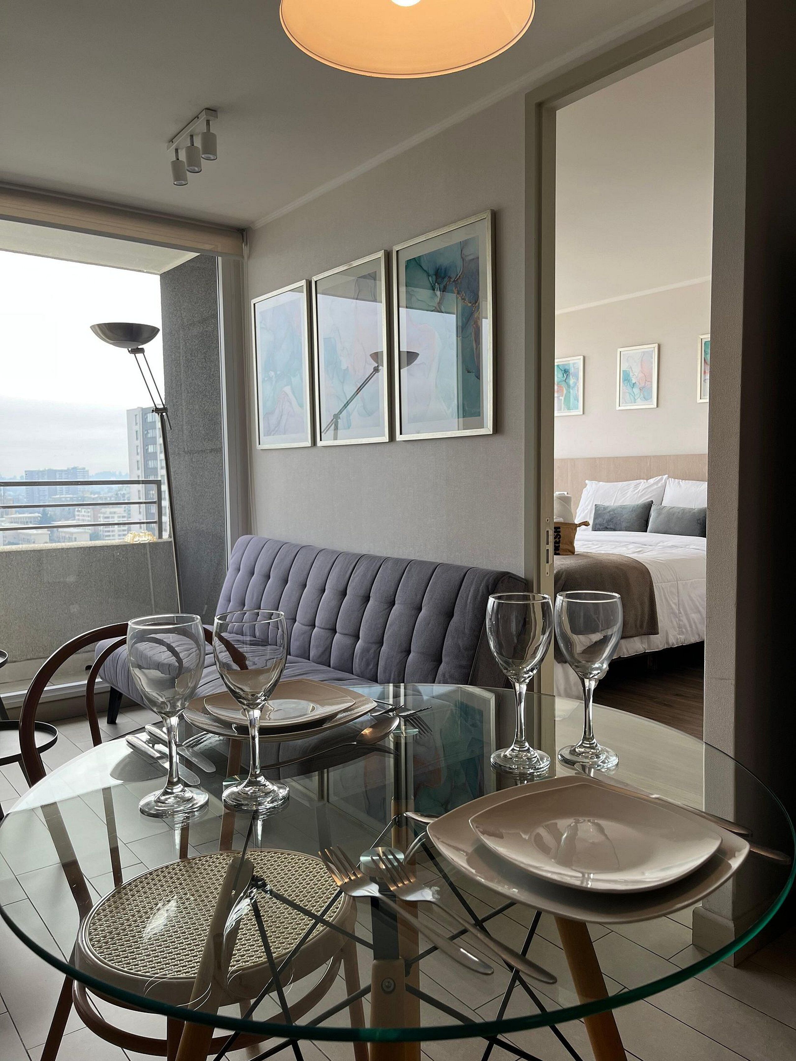 A modern dining area with a glass table, chairs, and a sofa. There are framed artworks on the wall and a bed in the background.