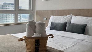 Ein Hotelzimmer mit großem Fenster, einem Bett mit weißen Leinentüchern, einem Korb mit Handtüchern, auf dem 'FRISCH' steht, und zwei abstrakten Bildern an der Wand.