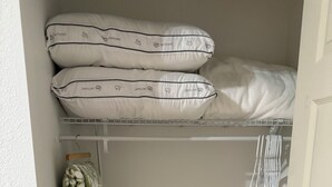 Un pequeño armario con un estante blanco que sostiene almohadas dobladas, una manta verde estampada colgada en la pared y un cesto de ropa rosa en el suelo.