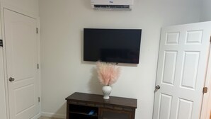 Una habitación con una puerta blanca, una pared blanca, un televisor de pantalla plana montado, una mesa consola de madera oscura con un florero de plumas rosas y un equipo de aire acondicionado blanco.