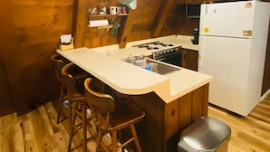 Une cuisine avec un intérieur en bois, un plan de travail blanc, une cuisinière à gaz, un évier, un réfrigérateur et des casseroles suspendues.