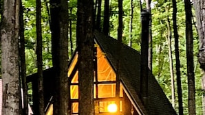 Une cabane nichée au cœur d’une forêt, dont la lumière chaleureuse de l’intérieur est visible à travers les fenêtres.